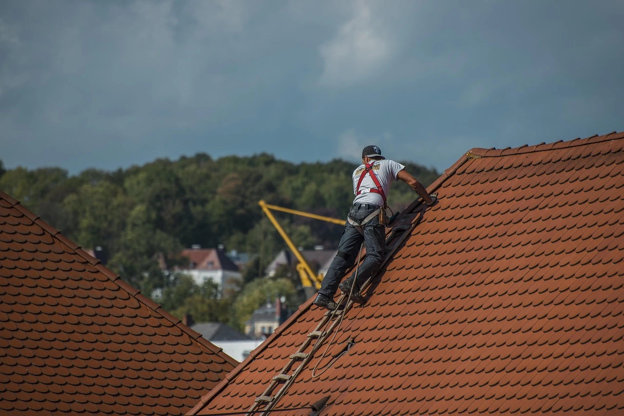 Roofer in a safety harness climbs a ladder on a steep tile roof under cloudy skies.
