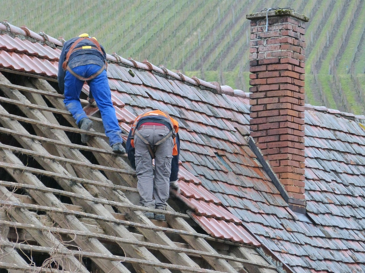 Two roofers replace tiles on a steep residential roof near a brick chimney.