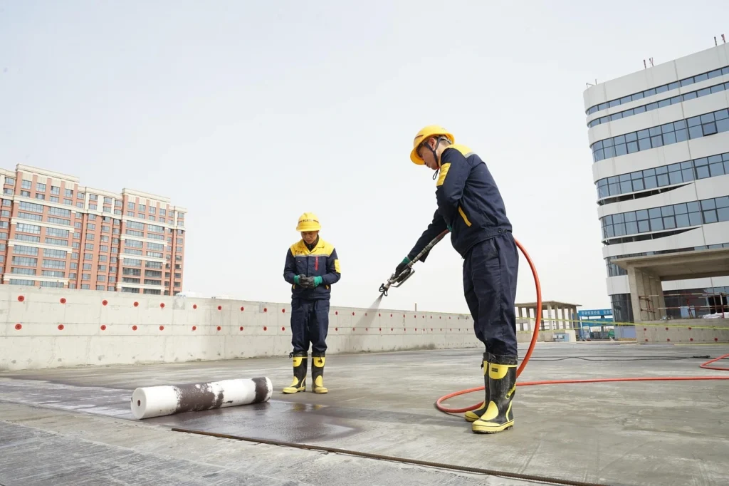 Two workers apply waterproofing material to a flat commercial roof with a spray hose and membrane roll.