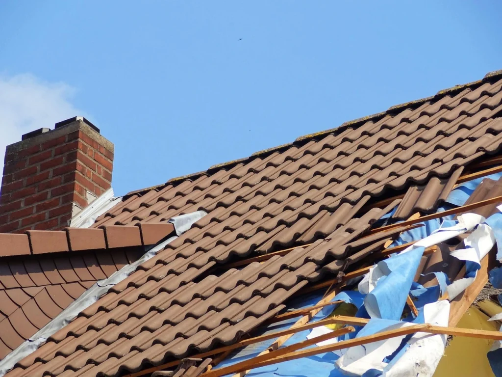 Storm-damaged tile roof with missing sections and exposed underlayment near a brick chimney.