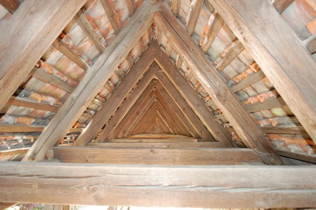 Attic view of wooden roof trusses and rafters forming a long triangular tunnel under clay tiles.
