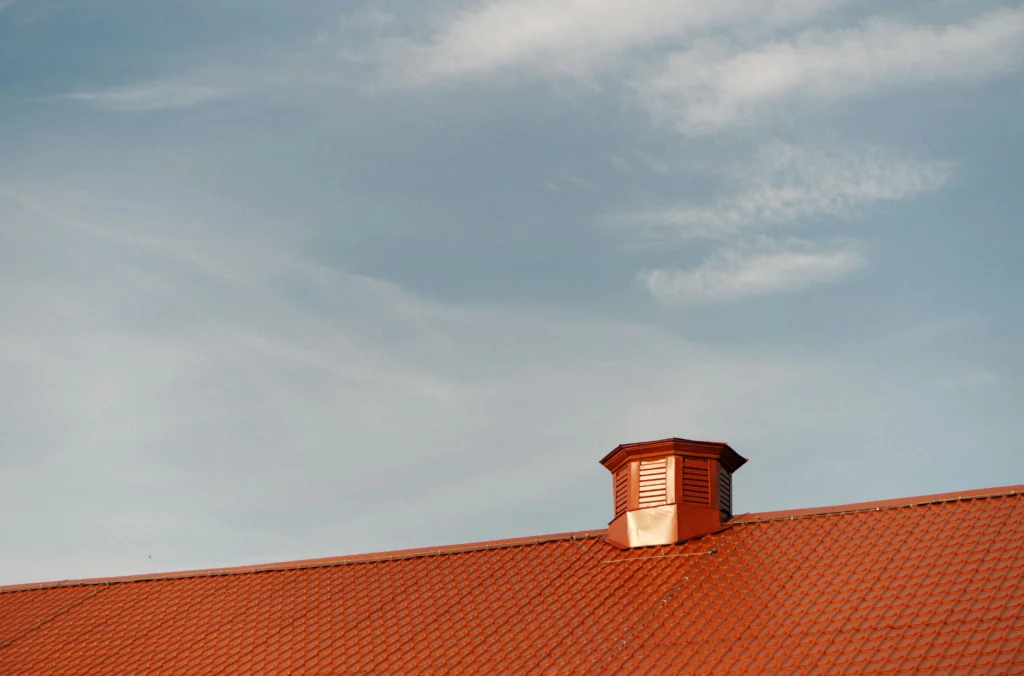 Red metal roof with a rooftop vent against a wide, partly cloudy sky.