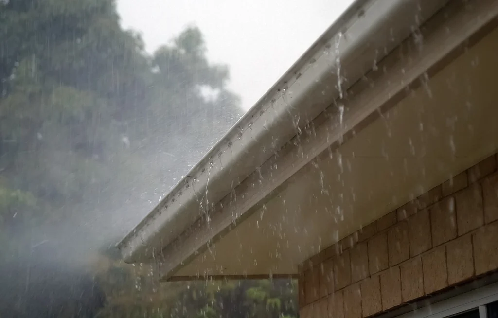 Heavy rain pours off a roof edge and gutter along the eaves of a brick home.