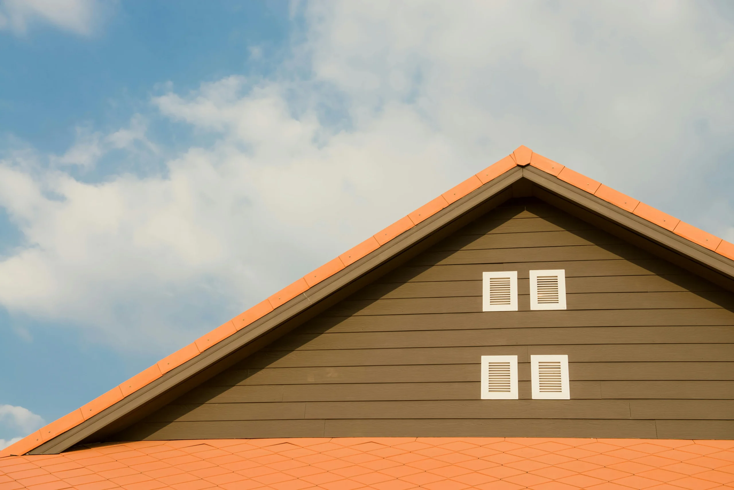 Gable roofline with orange tiles and two attic vents against a partly cloudy blue sky.